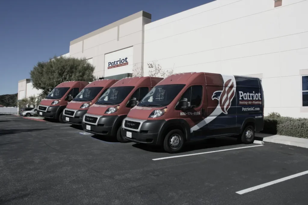 Four Patriot Heating & Air Conditioning vans are parked in a row outside a commercial building with the Patriot logo on the wall.