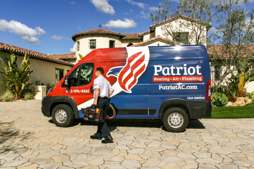 A technician walks toward a Patriot Heating, Air, Plumbing service van parked in the driveway of a residential home.